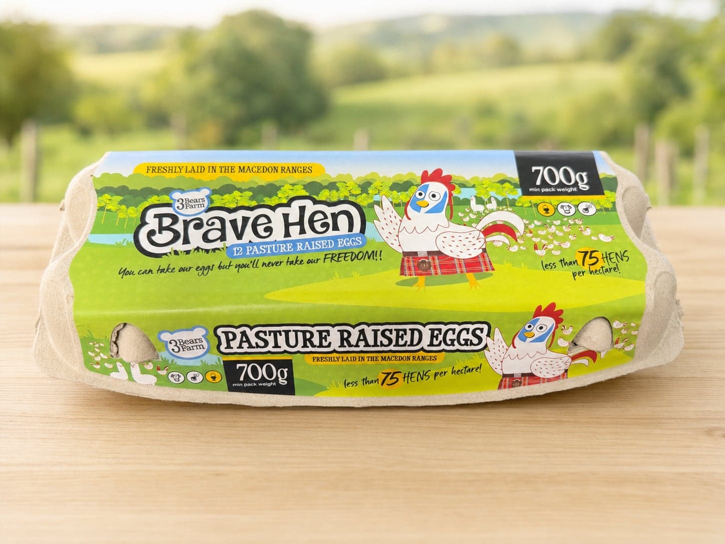 A closed 700 g dozen carton of Brave Hen pasture-raised eggs on a timber bench, green pasture behind